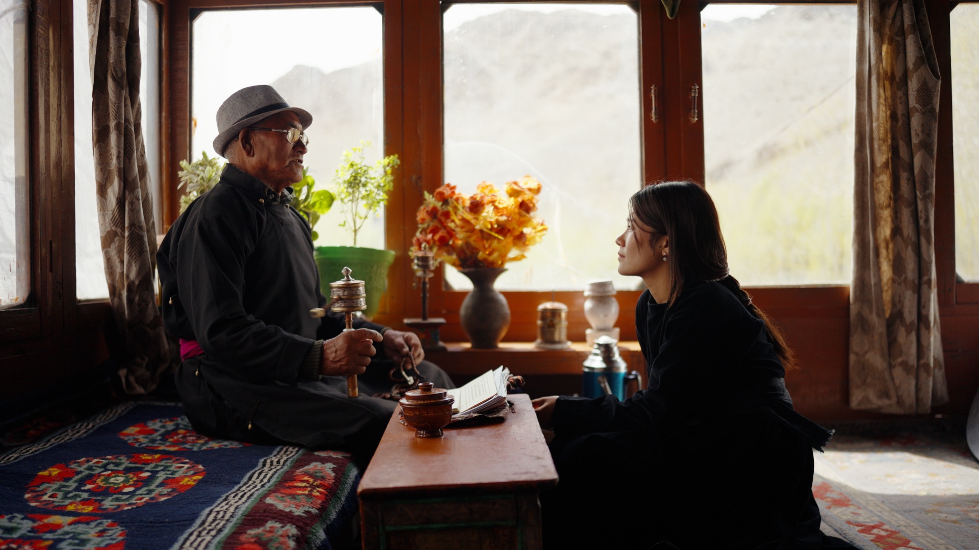 Stanzin sitting with a Ladakhi village elder holding a prayer wheel, warm interior light