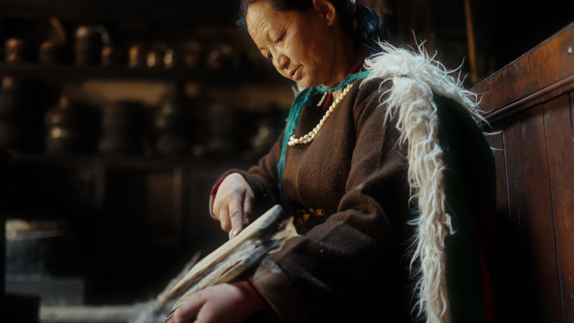 Ladakhi woman in traditional dress sitting in a warmly lit village room with traditional artifacts on shelves