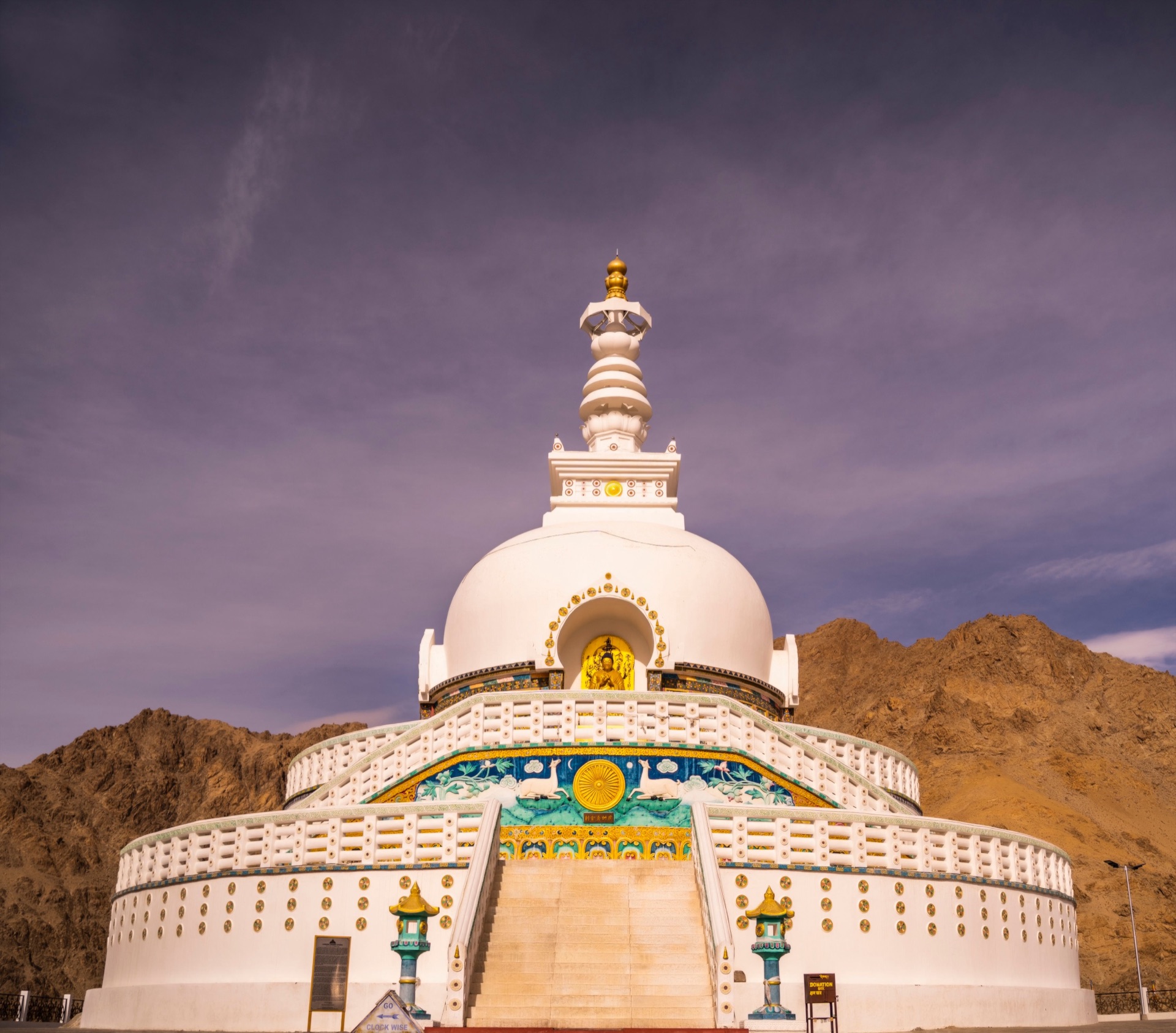 Shanti Stupa with colourful prayer flags against a dramatic sky, mountains behind