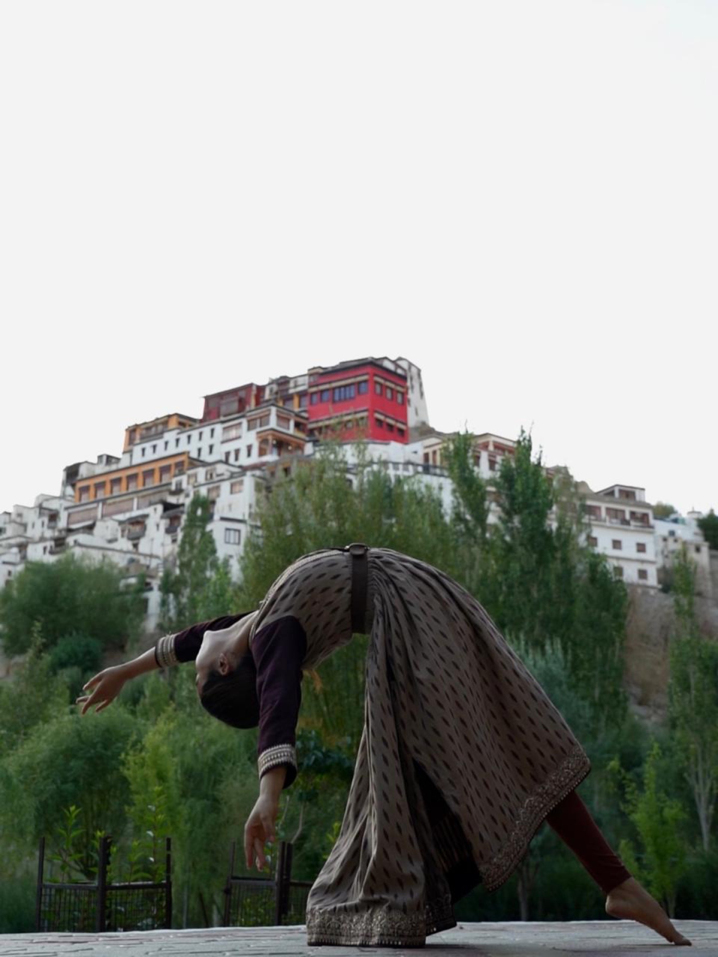 Yoga pose in front of Thiksey Monastery with green poplar trees and monastery walls behind
