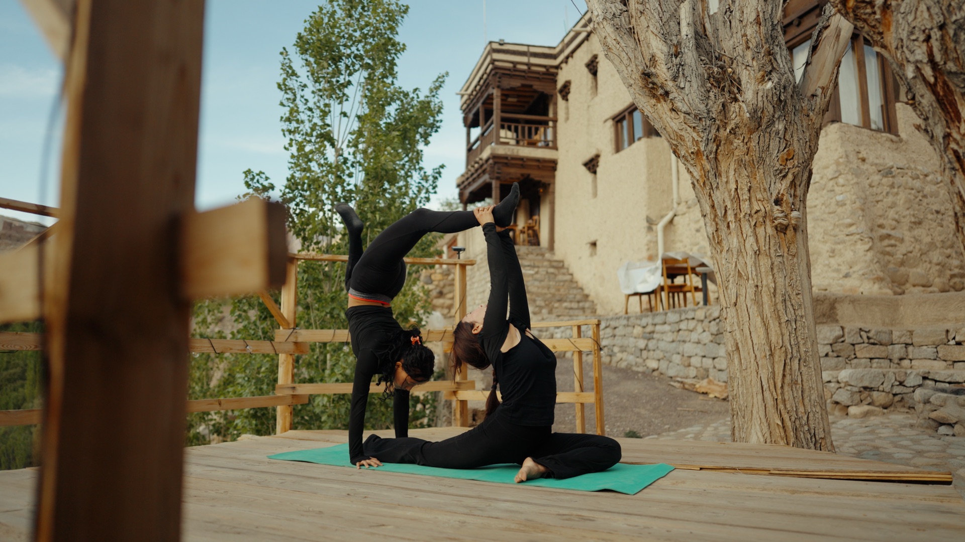 Two people doing partner yoga on a wooden deck with a traditional Ladakhi mud-brick building behind